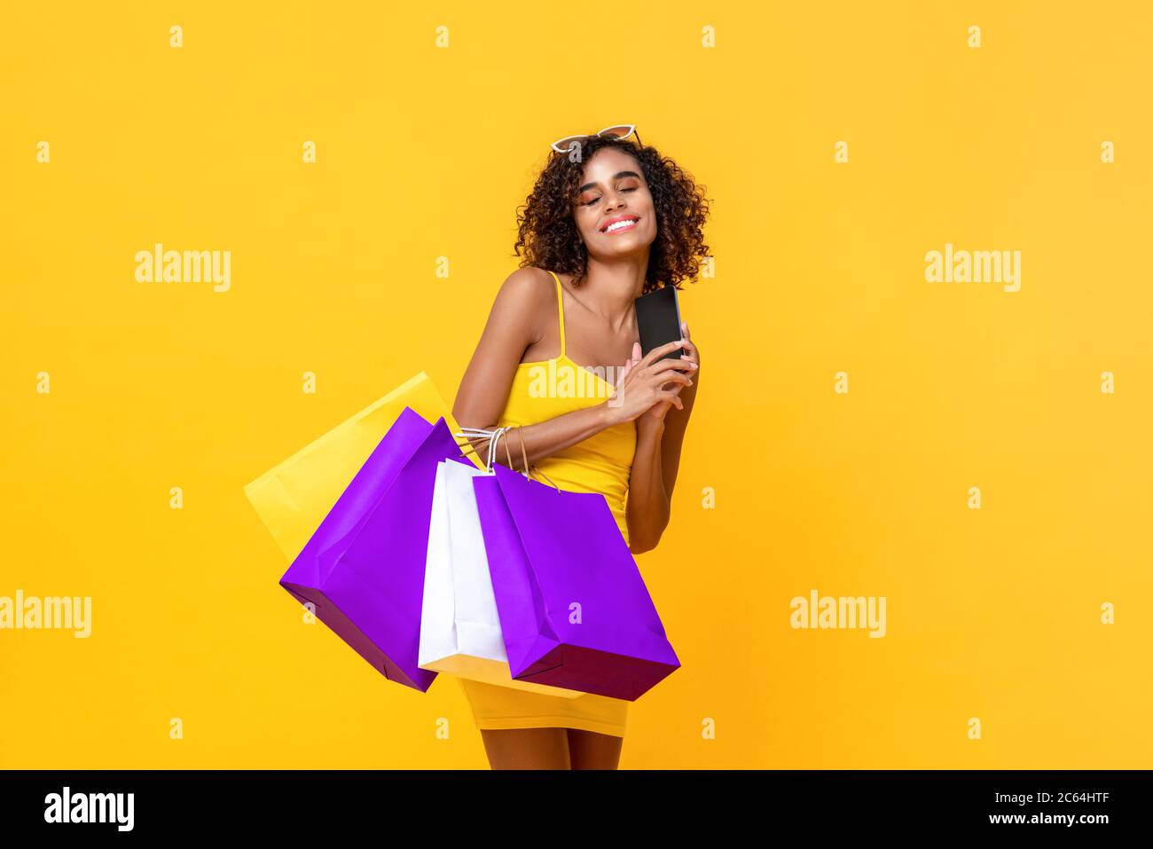 Portrait of satisfied attractive African American woman with eyes closed holding mobile phone and carrying shopping bags in isolated studio yellow bac Stock Photo