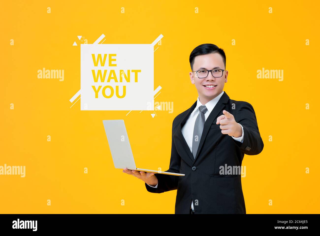 Job hiring concept portrait of smiling Asian businessman holding laptop computer while pointing hand on isolated yellow background with WE WANT YOU te Stock Photo
