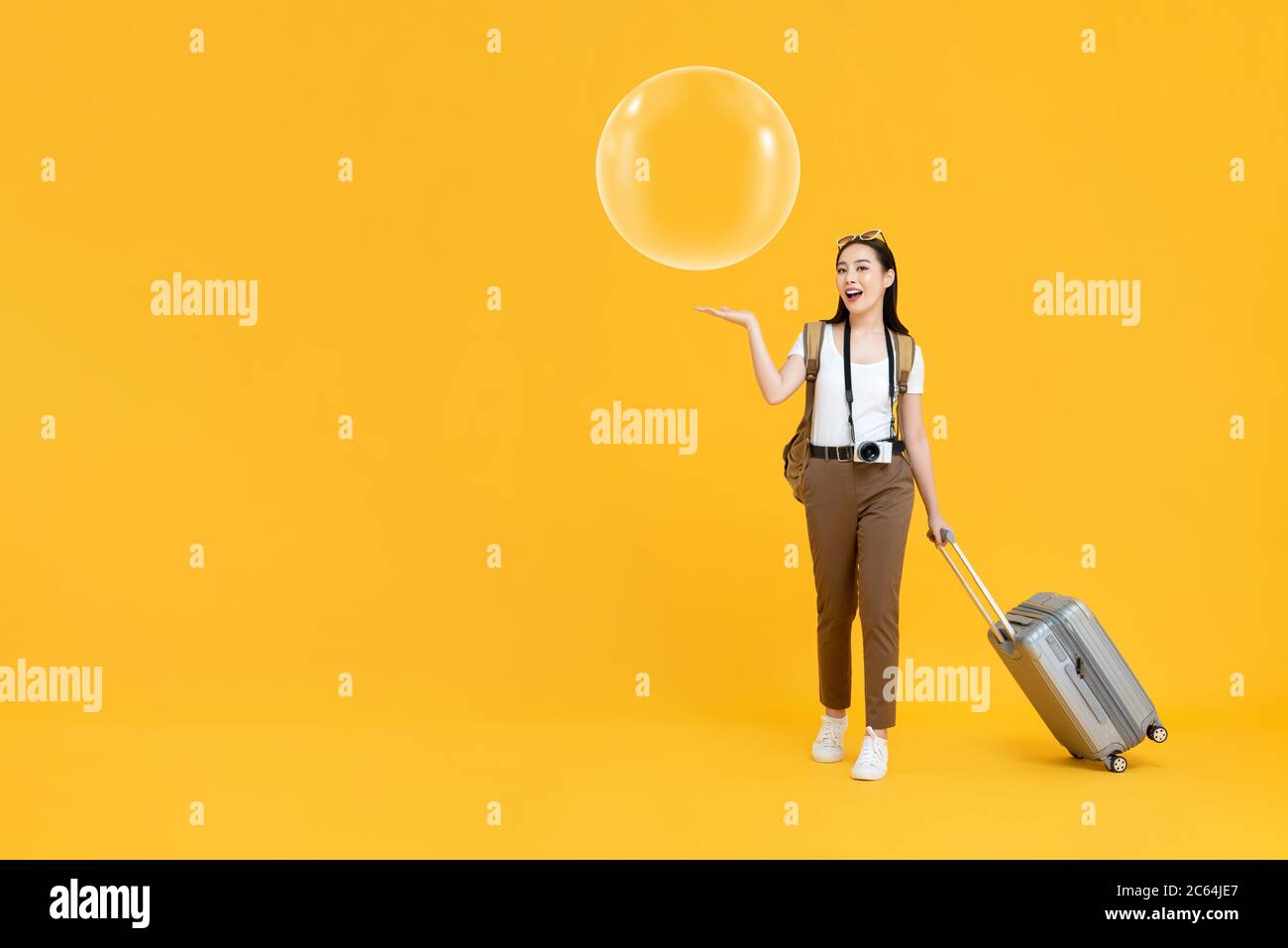 Full length portrait of  happy young Asian female tourist holding luggage with open palm showing bubble in isolated studio yellow background Stock Photo