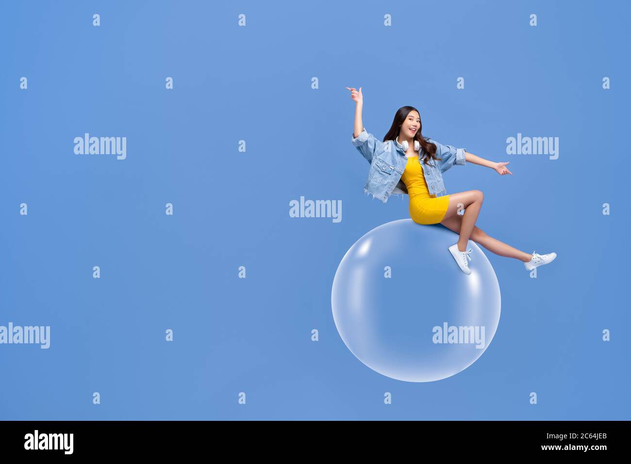 Fun portrait of smiling young Asian woman sitting on floating bubble with both fingers pointing sideways in isolated studio blue background Stock Photo