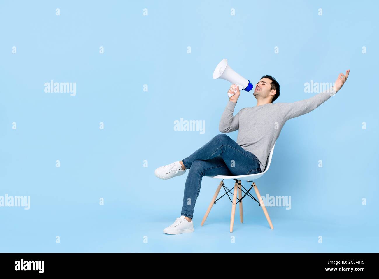 Portrait of young handsome Caucasian man sitting and cheerfully announcing on megaphone with one arm raised in isolated studio blue background Stock Photo