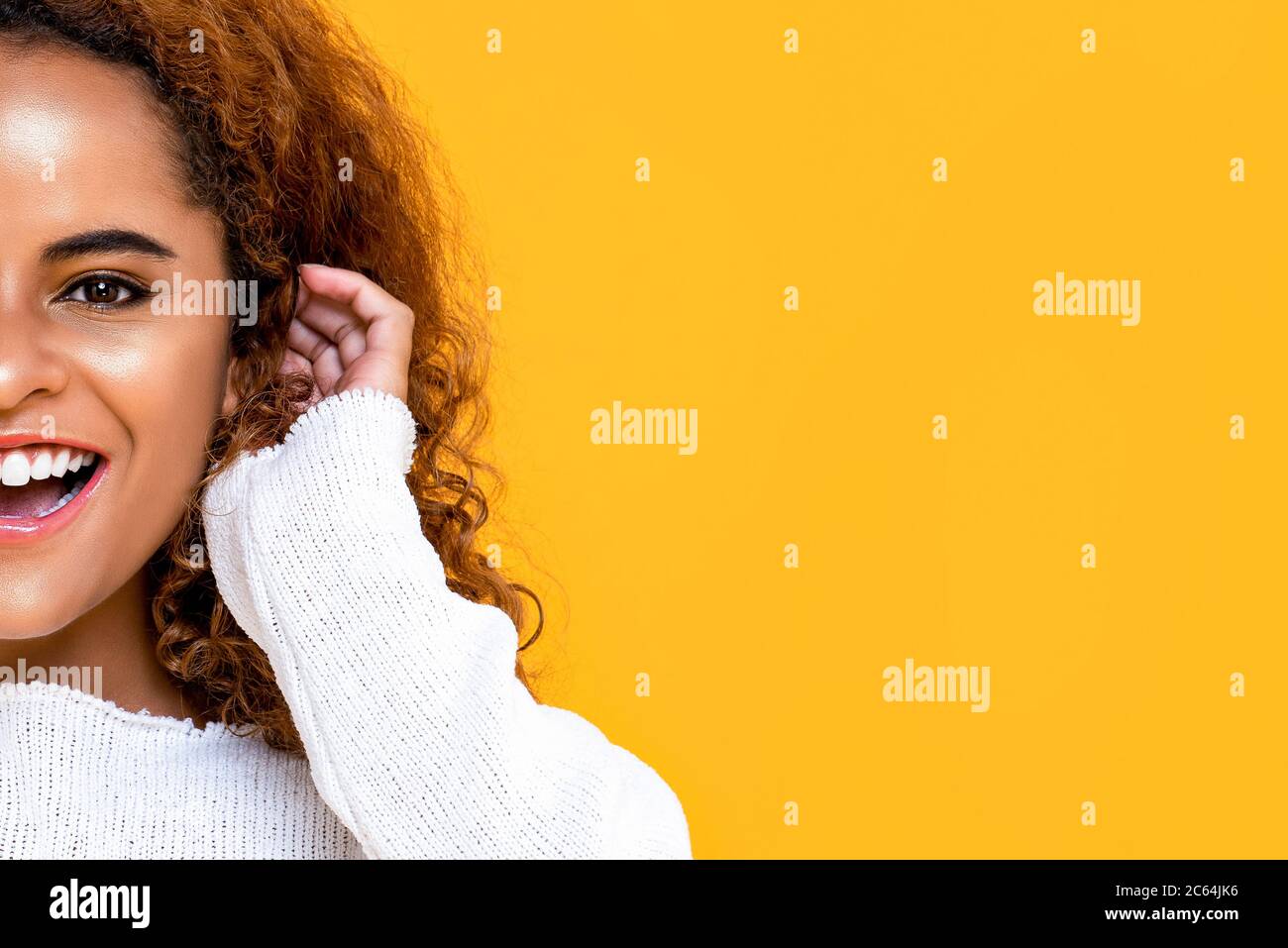 Half face close up portrait of young African American woman smiling while touching her ear in isolated yellow background with copy space Stock Photo