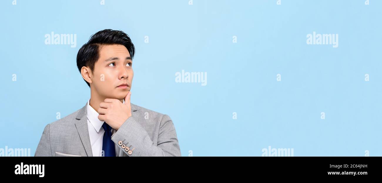 Panoramic portrait of serious young Asian businessman thinking with hand on chin isolated blue background with copy space Stock Photo