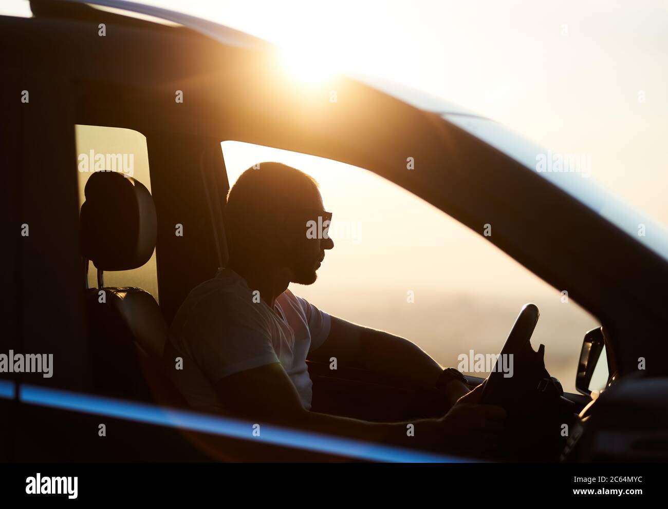 Silhouette of a young man sitting in his car, wearing sunglasses and looking at the sunset, hand on the helm, bright sun on the car roof, close up, side view Stock Photo