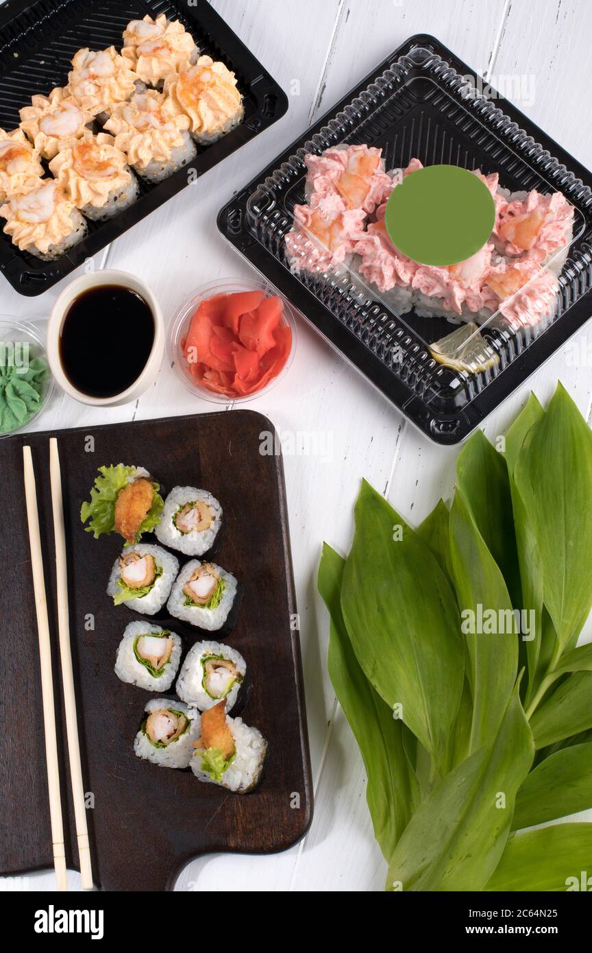 Sushi rolls set served on dark wooden board and plastic boxes. Restaurant food delivery in take away boxes. Gray background with green leafs Stock Photo