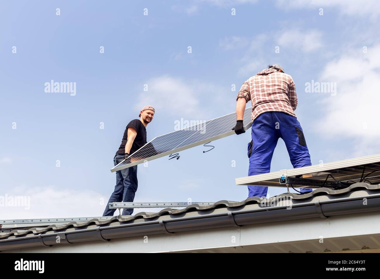 Happy electrical engineers mans are holding solar panel on roof against blue sky. Development sun alternative energy technology. Ecological concept. Stock Photo