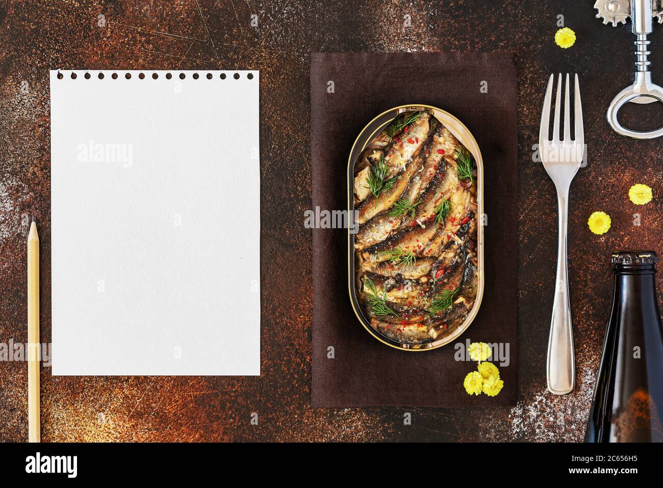 Open tin of Baltic sprats in oil with spices with a fork and a bottle of dark beer with a sheet of white writing paper Stock Photo