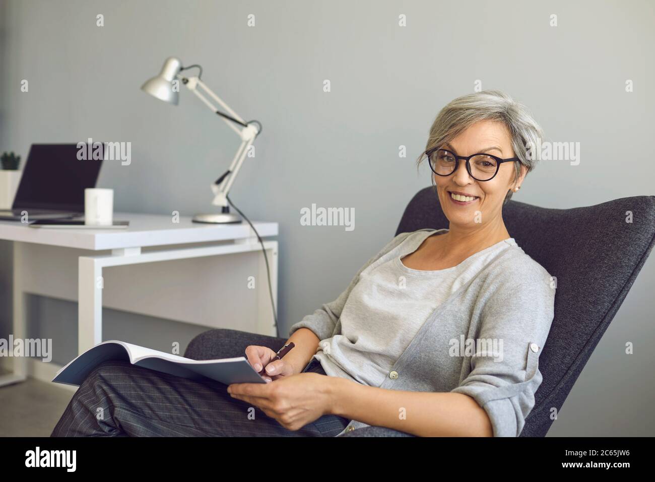 Happy senier woman in glasses writes a text in a textbook sitting in a chair at home. Work and education for people of age. Stock Photo