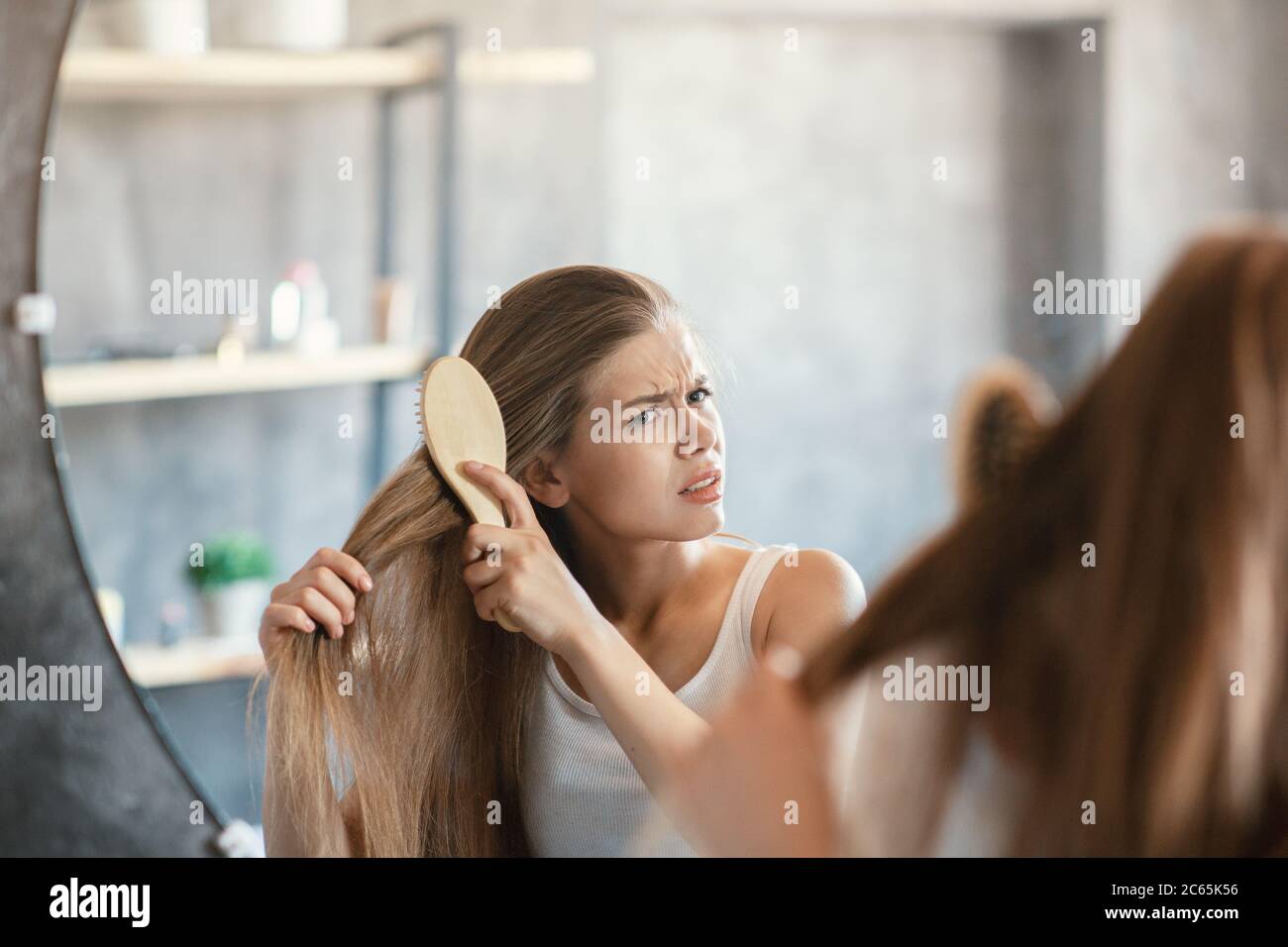 Frustrated young woman trying to brush her tangled hair in front of mirror in bathroom Stock Photo