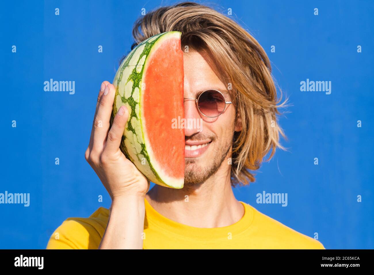 Attractive young man with long blond hair with half a watermelon covering his face on a plain blue background. Summer, sun, heat, fruit, vacation conc Stock Photo