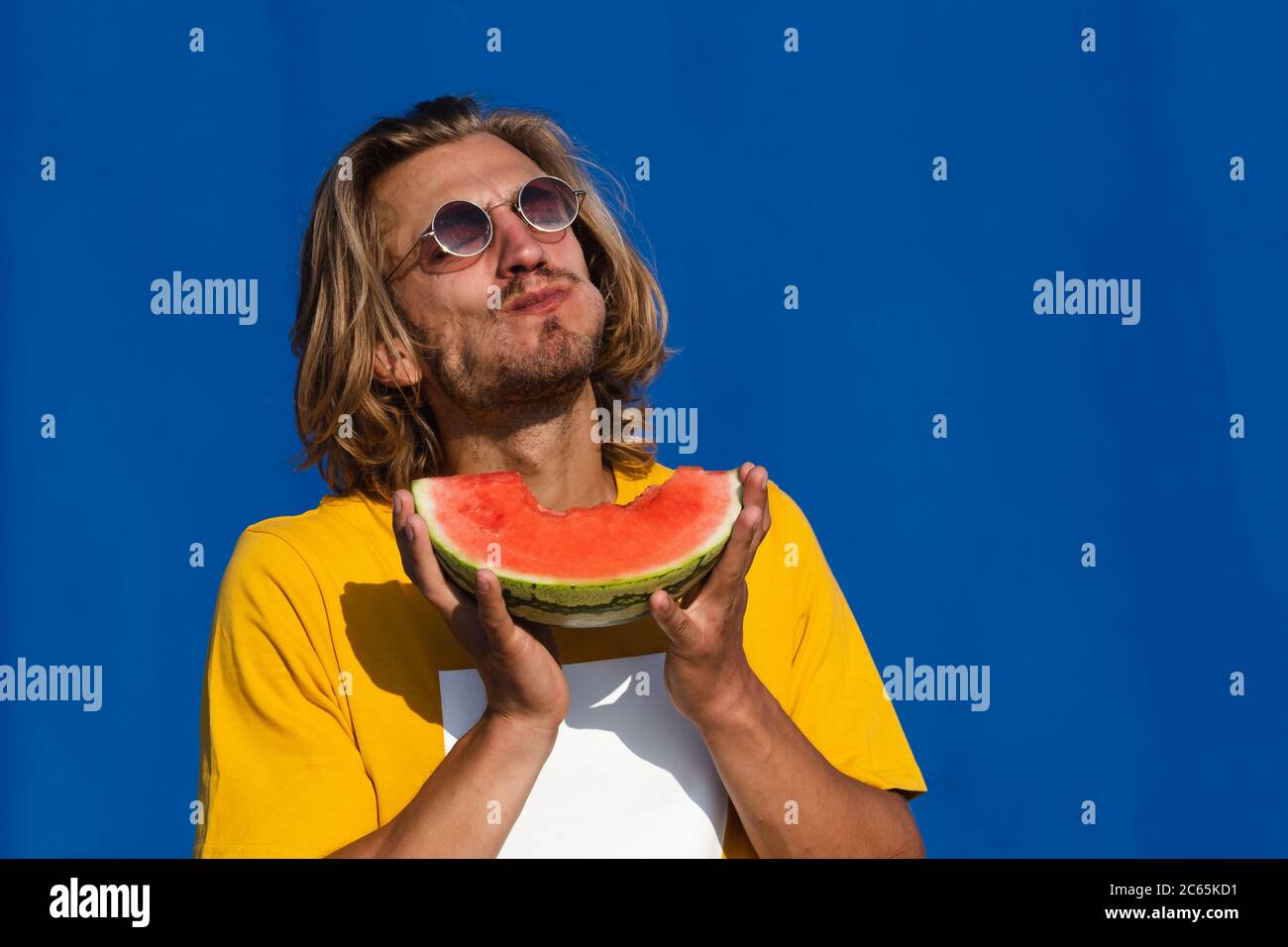 Attractive young man with long blond hair eating half a watermelon and enjoying on a plain blue background. Concept summer, sun, heat, fruit, holidays Stock Photo