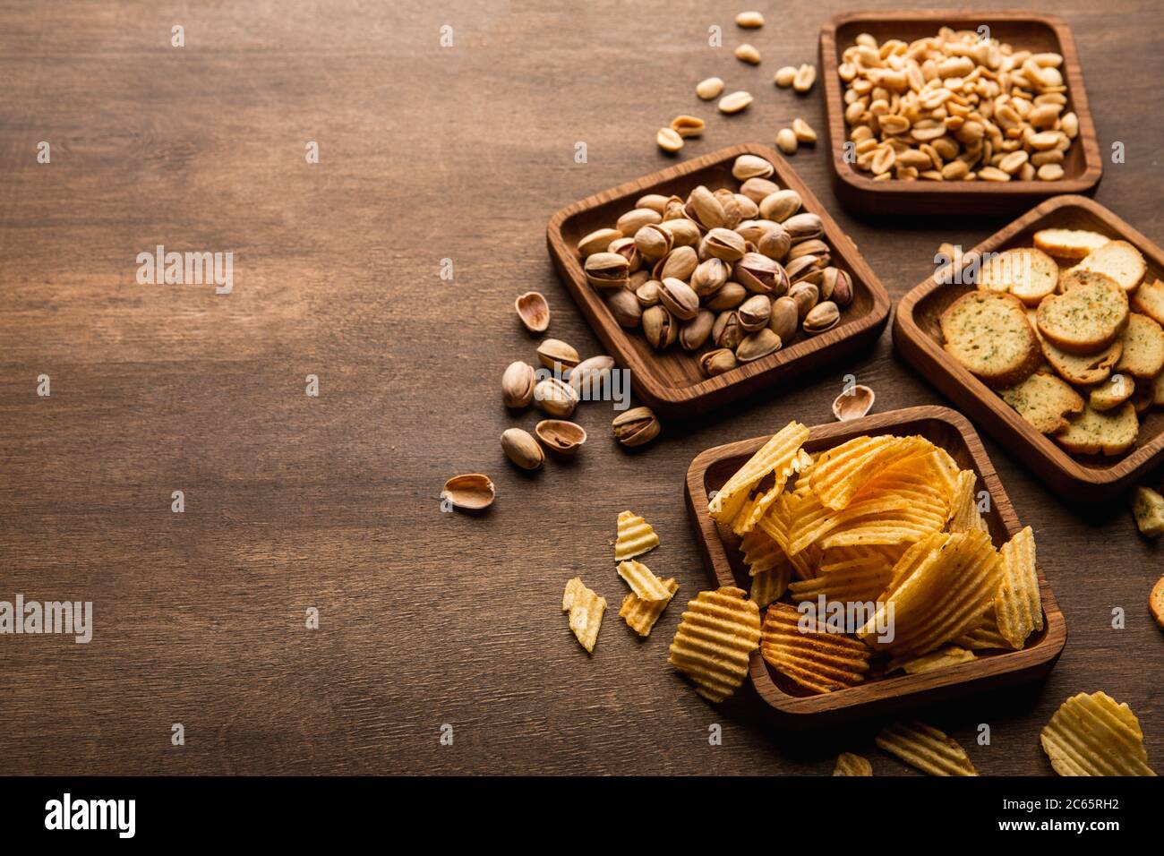 Crispy snacks. Rusks, peanuts pistachios and chips in square wooden plates on brown table Stock Photo