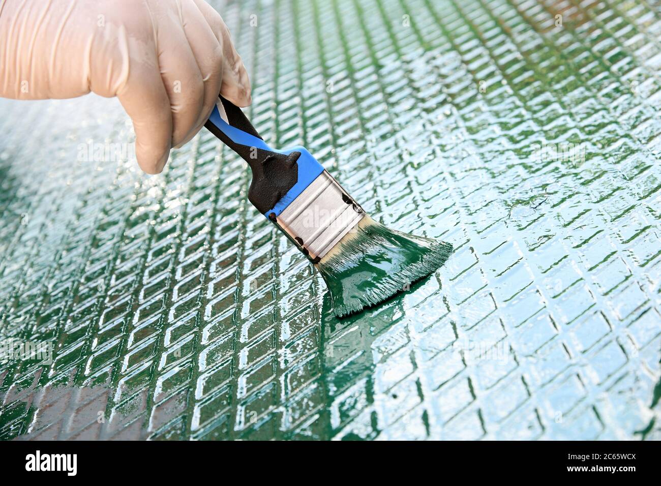 Person refreshing the green paint on a metal grid with diamond pattern in a close up view of a gloved hand and paintbrush Stock Photo