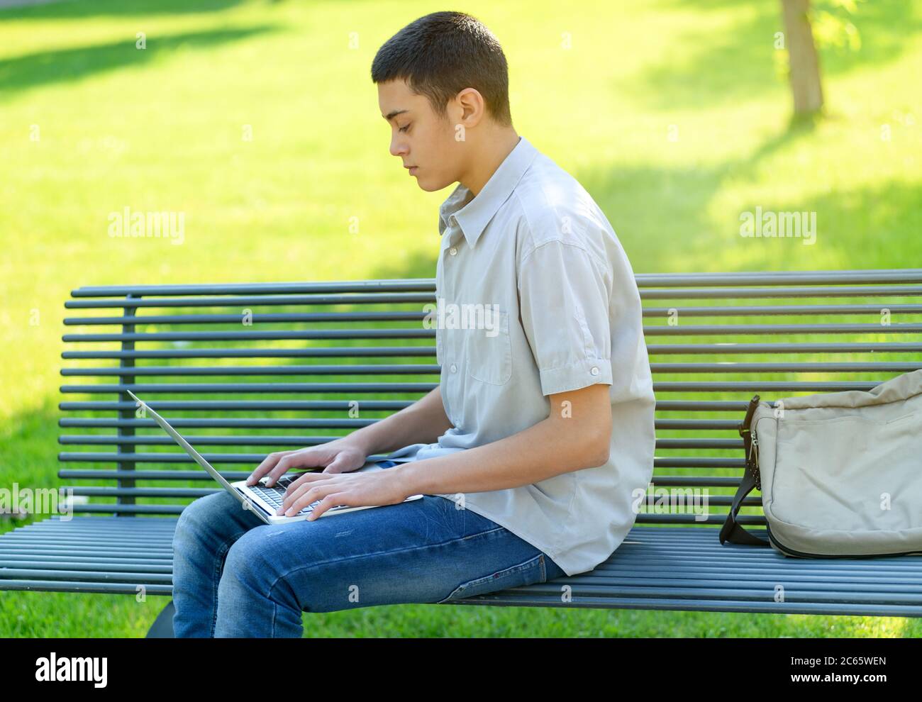 Young teenage boy using his laptop in the park as he sits on a bench typing with it balanced on his lap on a sunny summer day against a green grass ba Stock Photo