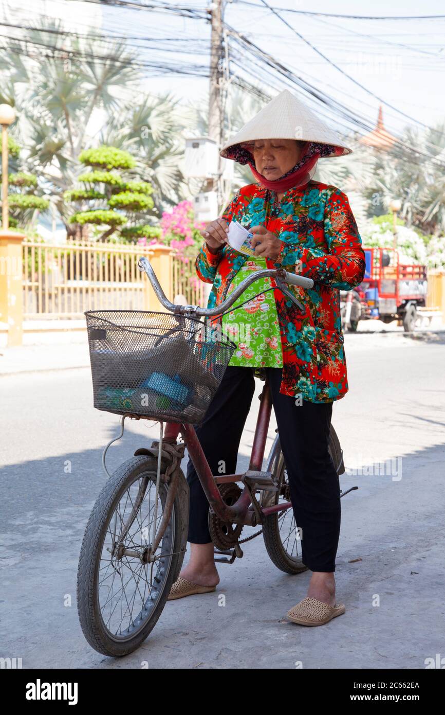 Local Vietnamese woman sitting on her bicycle with a basket kerbside whilst out shopping checking some tickets in street near My Tho City, Vietnam Stock Photo