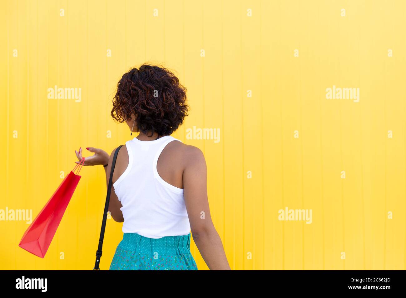 Back view of woman walking with a shopping bag next to a yellow wall. Space for text. Concept of shopping, gifts and sales. Stock Photo