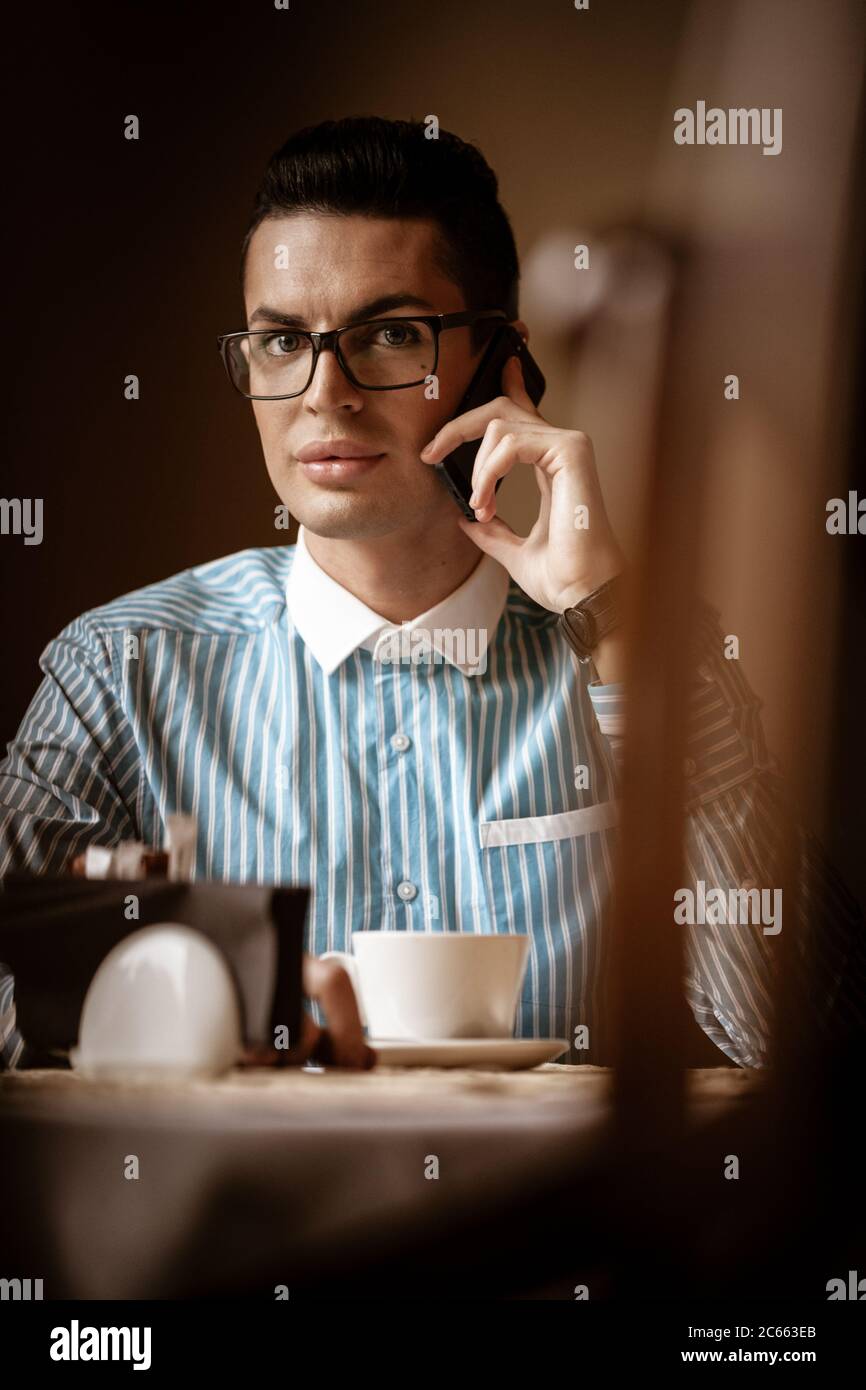 LGBTQ community lifestyle concept. Young homosexual man sits at the table in old-fashioned city cafe. Handsome gay male businessman talks on a phone Stock Photo
