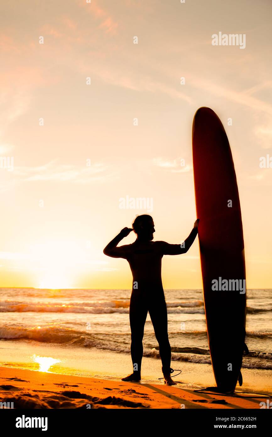 Male surfer silhouette with yellow longboard on a ocean coast at sunset. Water sport adventure camp and extreme swim on summer vacation. Stock Photo