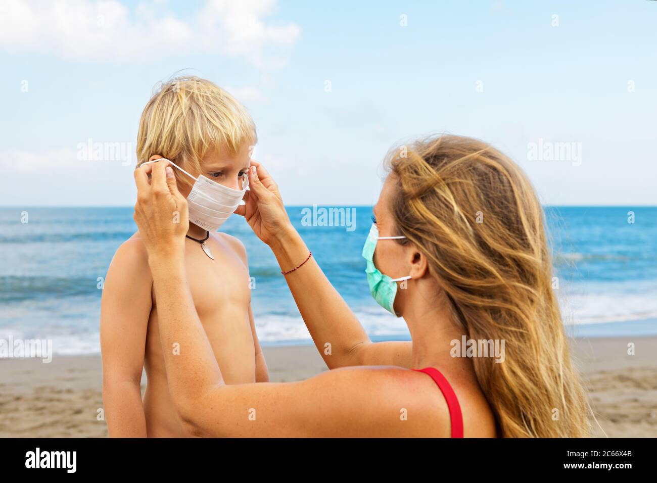 Mother put medical mask on child on sea beach. New rules to wear cloth face covering at public places. Cancelled cruise, tour due coronavirus COVID 19 Stock Photo