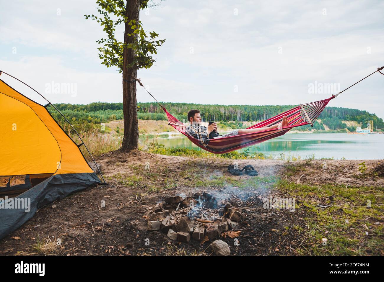 man laying on hammock at lake beach near camp fire Stock Photo