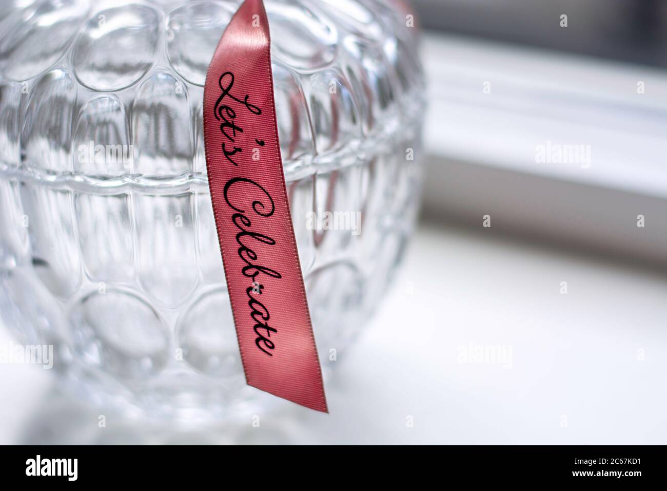Decorative retro glass jar container with a pattern with a pink ribbon with text Let's Celebrate on a white windowsill. Stock Photo