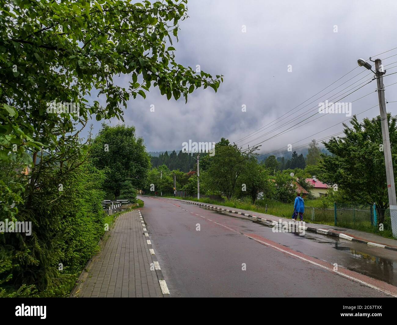 Woman pedestrian in rainwear walking along the country road in carpathian mountains. Cloudy weather with dark storm clouds. Stock Photo