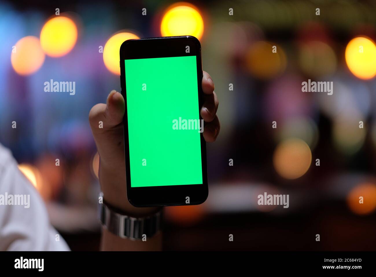close up one hand showing a green screen smartphone with colorful defocused lights background. Bar restaurant concept Stock Photo