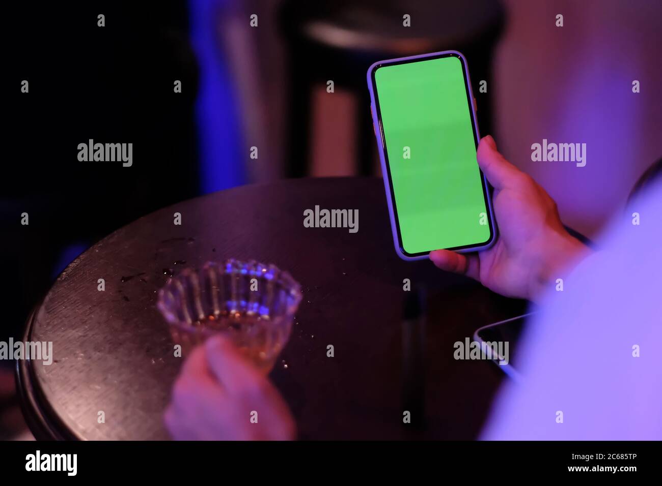 over shoulder of man holding green screen phone at bar. blurred wooden table and cocktail glass. Stock Photo