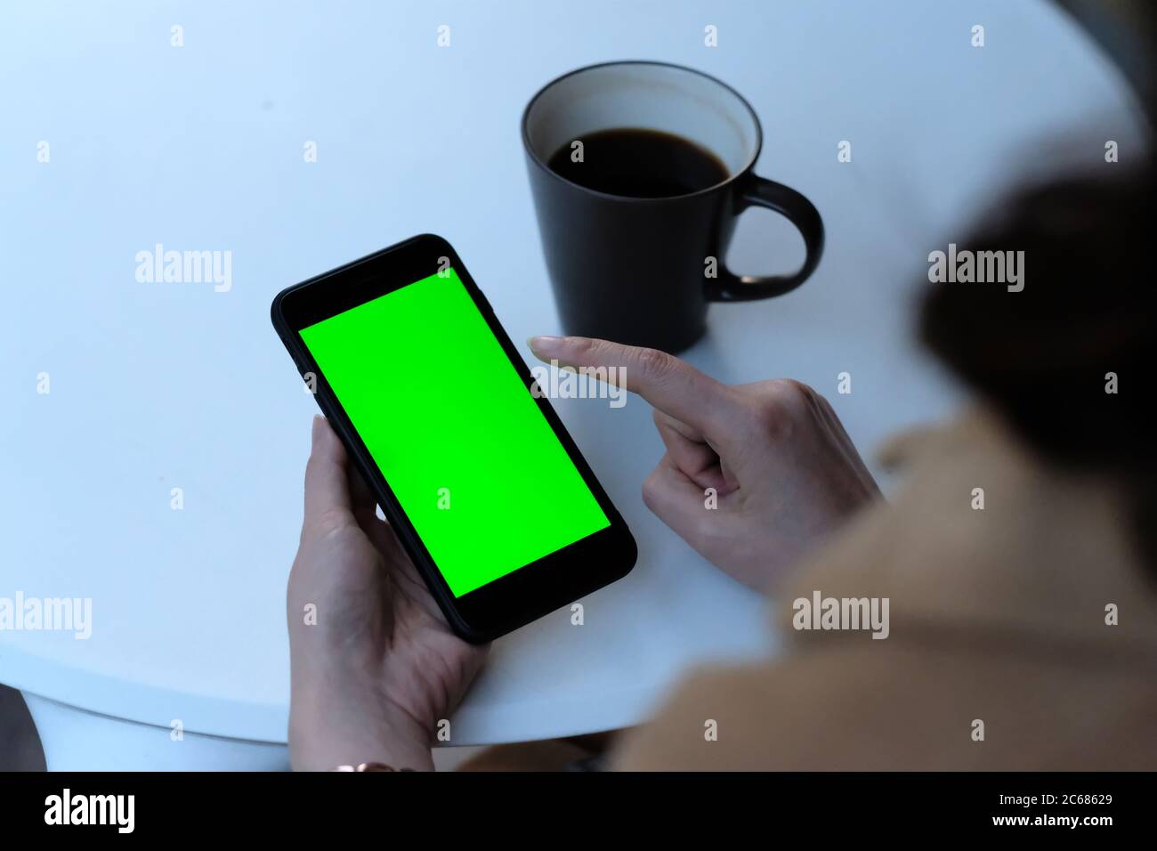 over should of woman tapping green screen smartphone at cafe. A cup of coffee on white table. Blur background Stock Photo