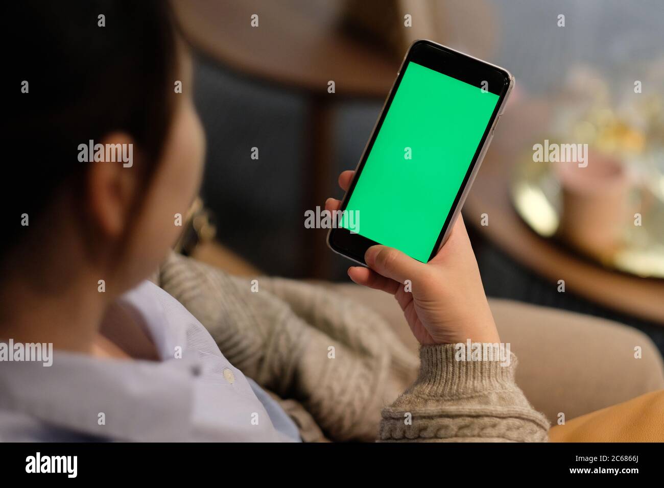 over shoulder of one young woman holding a green screen smartphone in living room at night. blur background Stock Photo