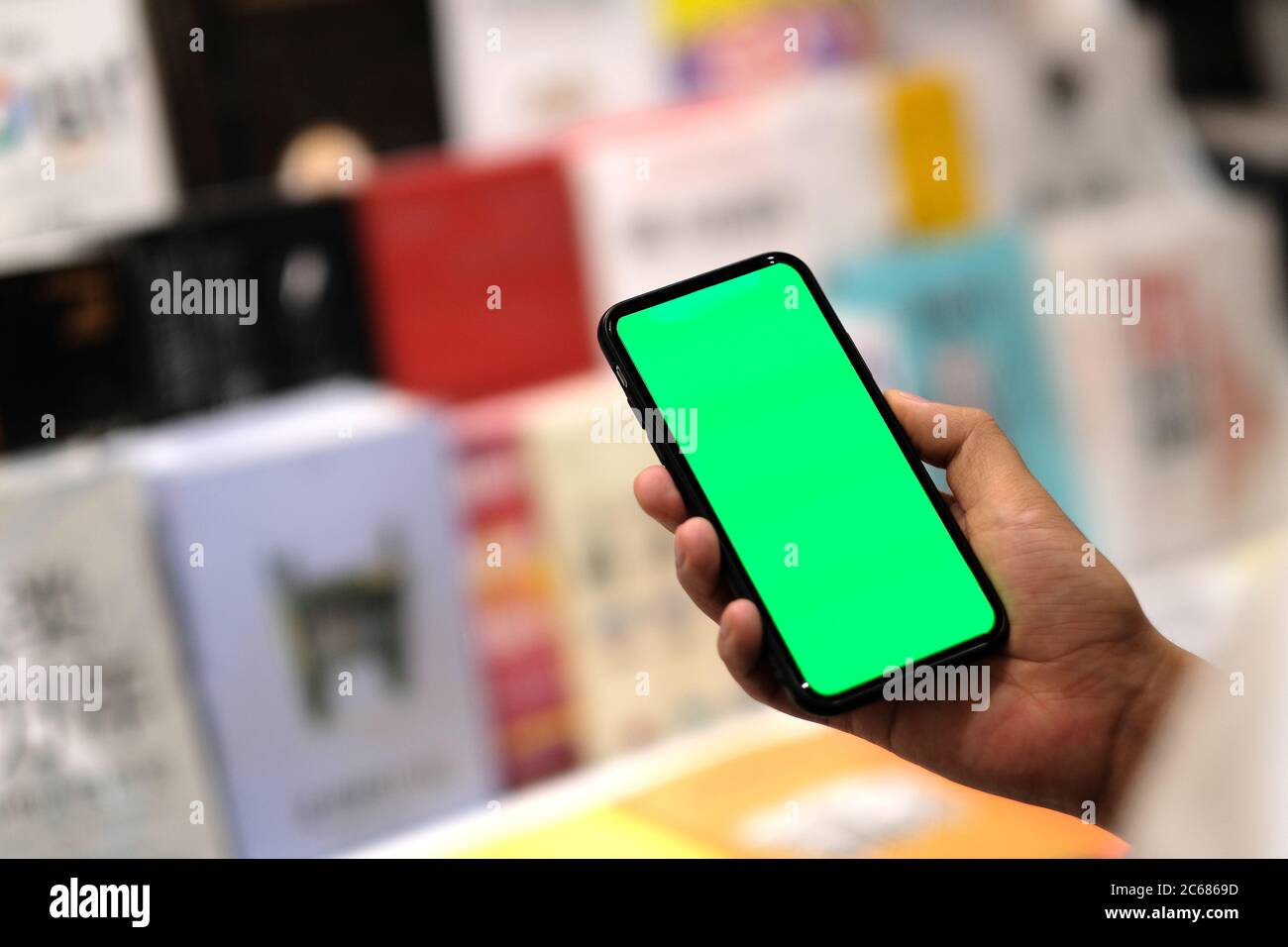over shoulder view of man holding green screen phone in book store. blurred books as background Stock Photo