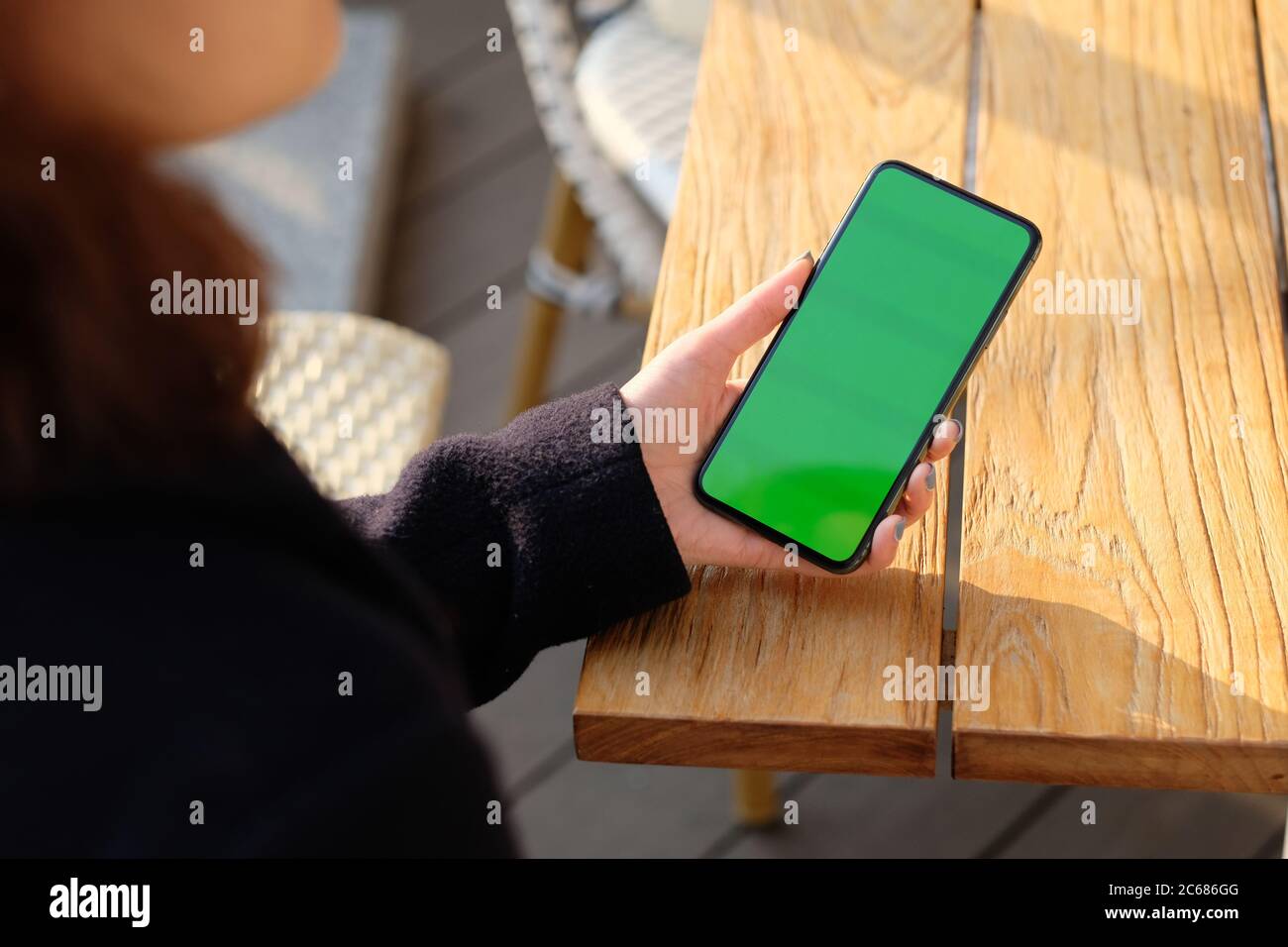 over shoulder of one woman holding green screen phone on wooden table under sunshine. blur background Stock Photo
