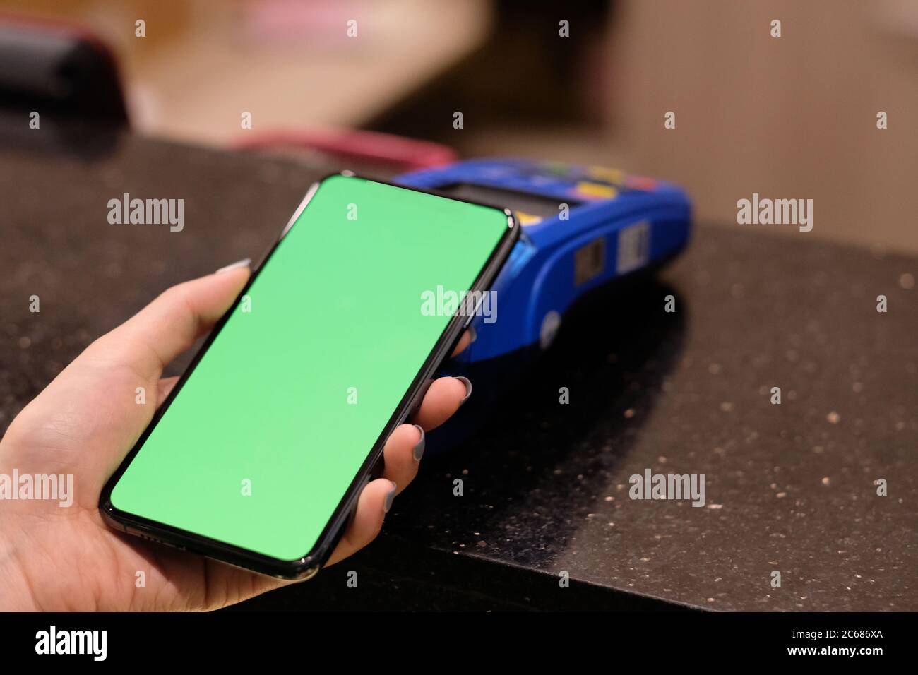 one hand holding green screen phone close to POS terminal on counter. Blur background. Contactless payment Stock Photo