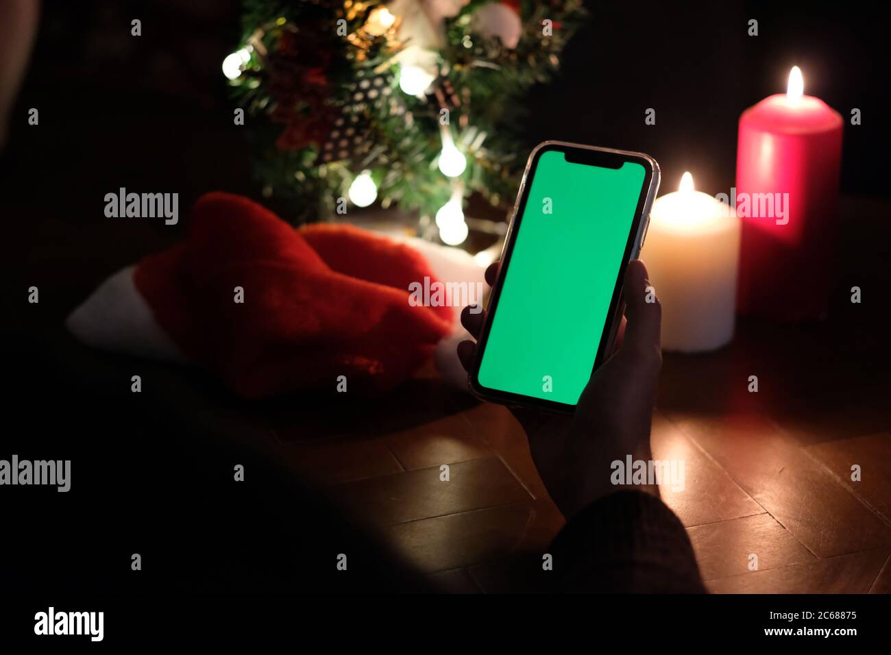 over shoulder of one hand holding green screen smartphone. Blurred christmas tree, hat and candle lights. black background Stock Photo