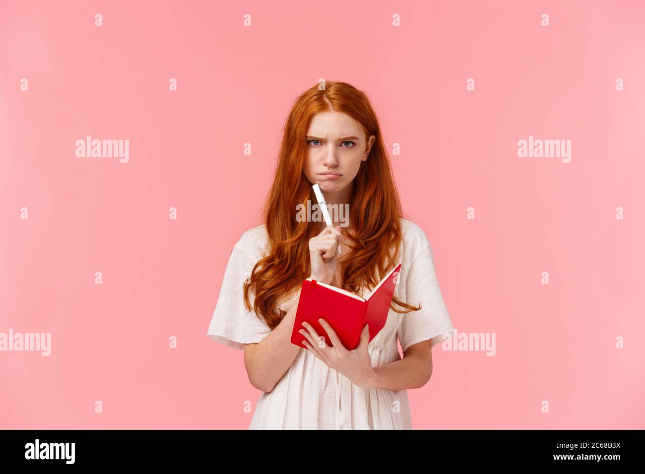 Serious-looking grumpy redhead woman frowning as thinking, pouting troubled, solving serious riddle, holding red notebook and touching chin with pen Stock Photo