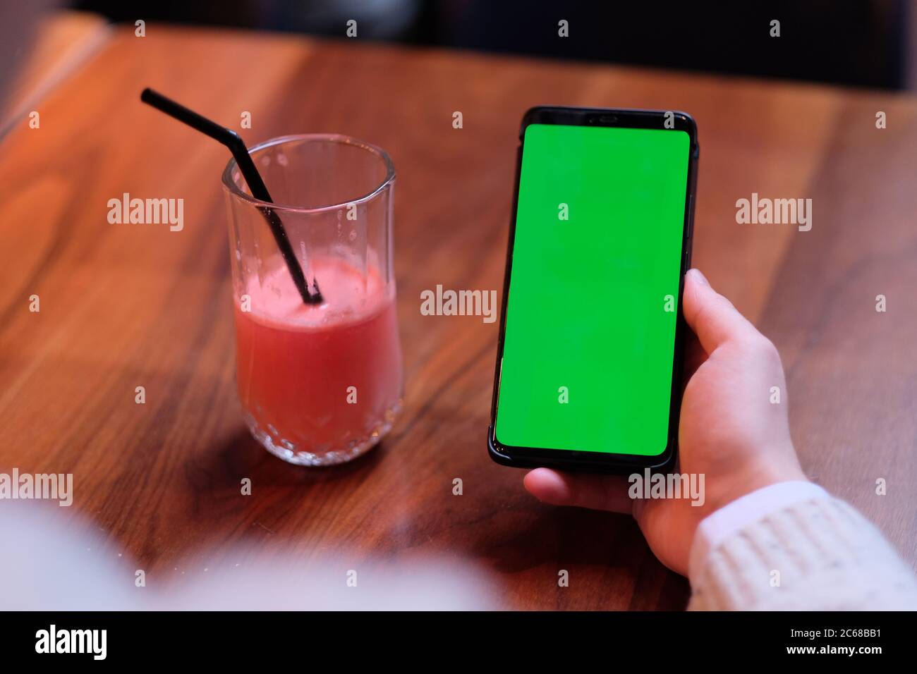over shoulder of man holding green screen phone with a glass of fruit juice on wooden table. Blur background Stock Photo