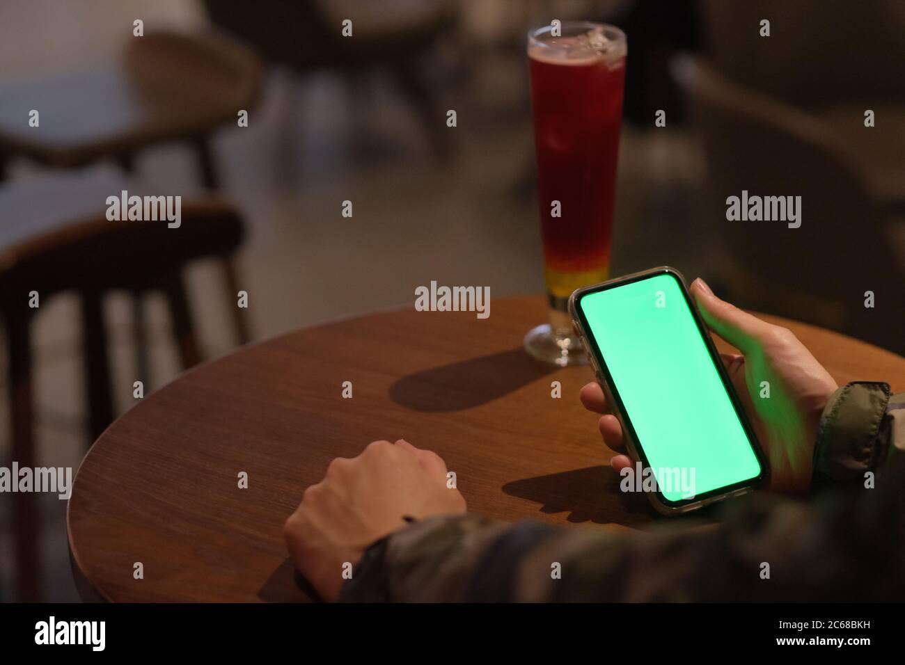 Over shoulder of man holding green screen smart phone in the bar at night. A glass of cocktail on woode table. Blur background Stock Photo