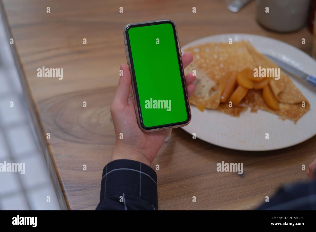 over shoulder of hand holding green screen smartphone. Blur food in plate on wooden table. Stock Photo