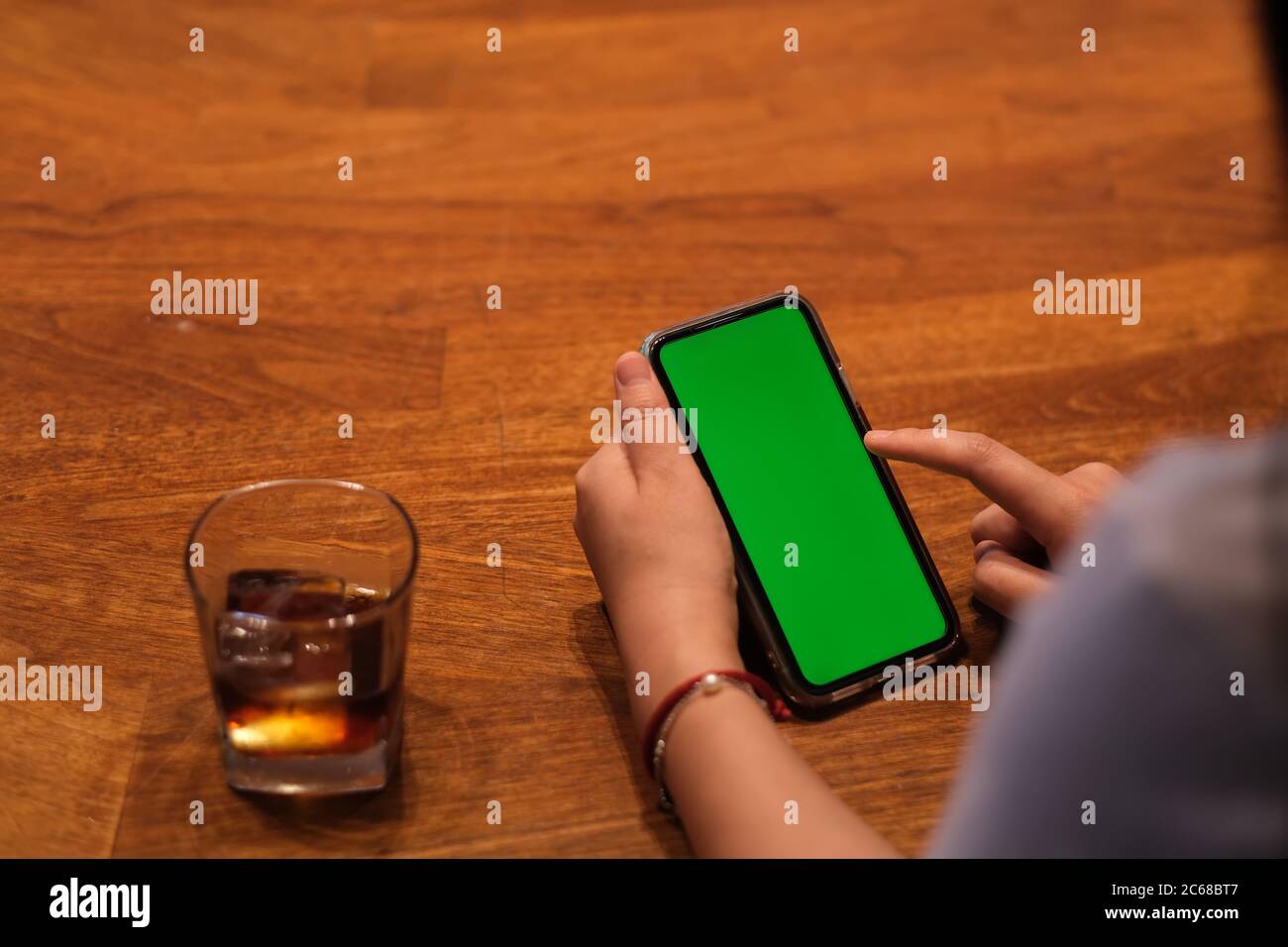 over shoulder of people tapping green screen smart phone. a glass of iced drink on wooden table Stock Photo