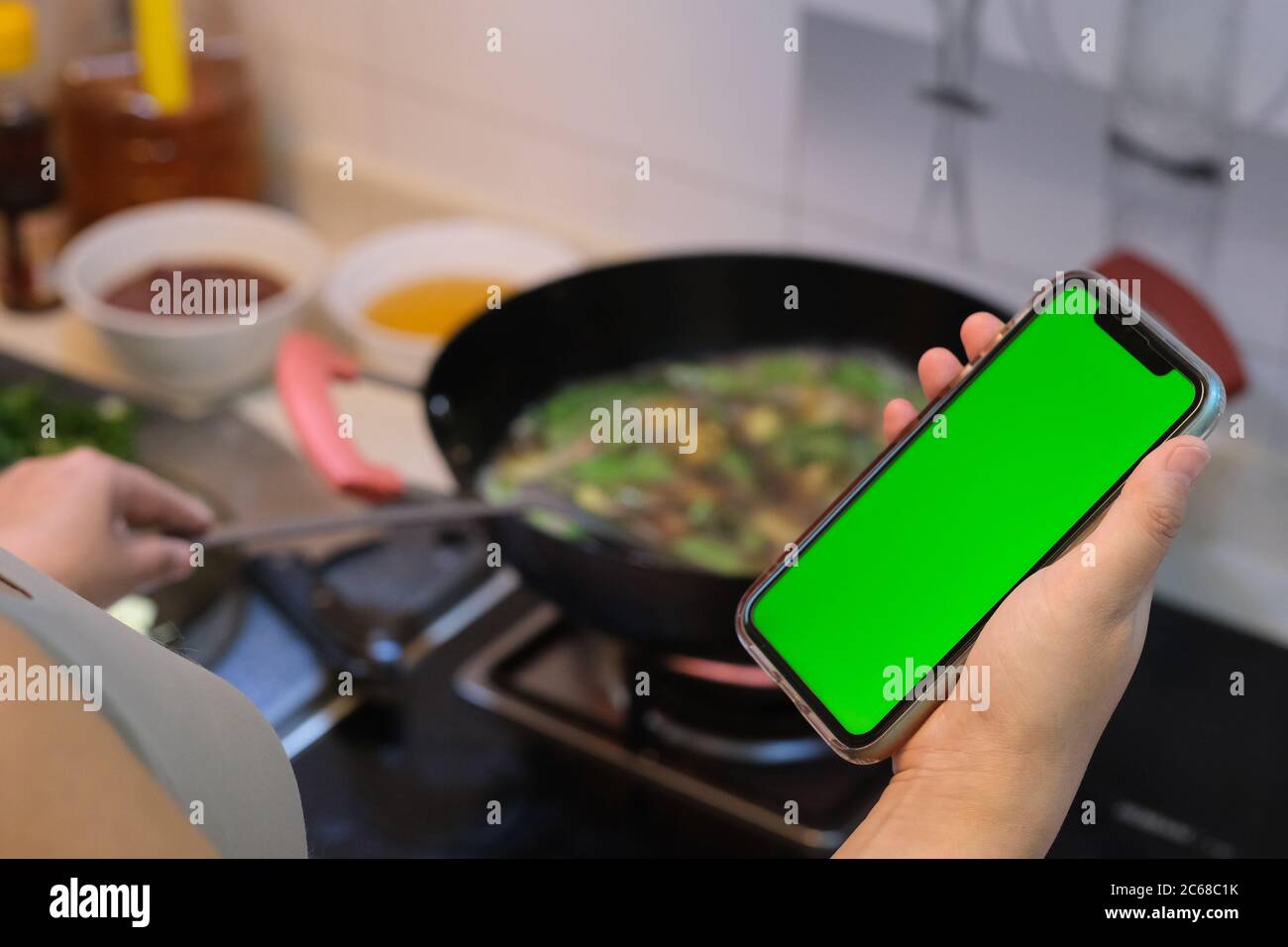 over shoulder of woman using green screen smartphone while cooking in kitchen. blur background Stock Photo