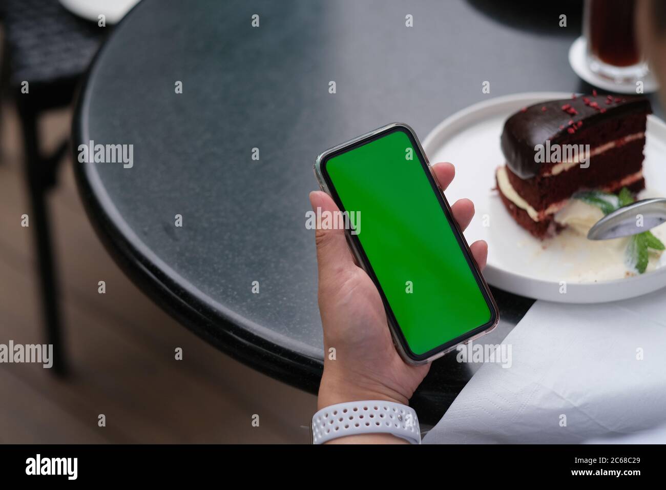 over shoulder of people holding green screen smartphone. blurred chocolate cake on black table. Stock Photo