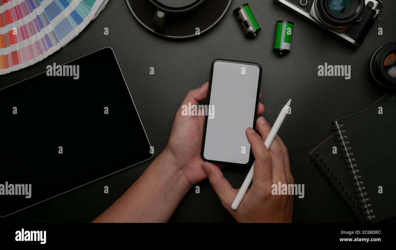 Top view of male designer holding mock-up smartphone and working on digital tablet and supplies on dark office desk Stock Photo