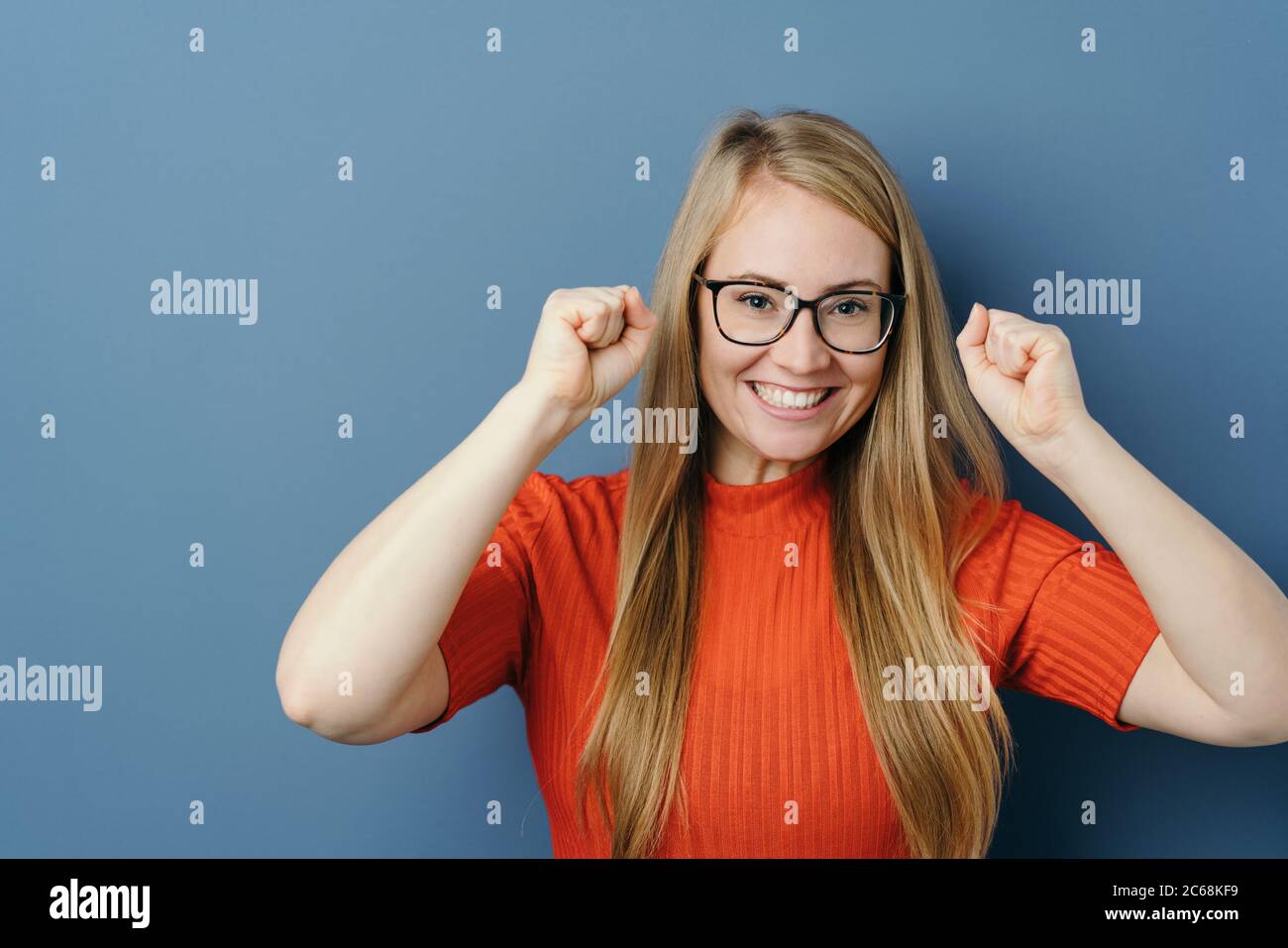 Elated young woman cheering and punching the air with her fists with a beaming smile after receiving good news of a personal success over a blue studi Stock Photo
