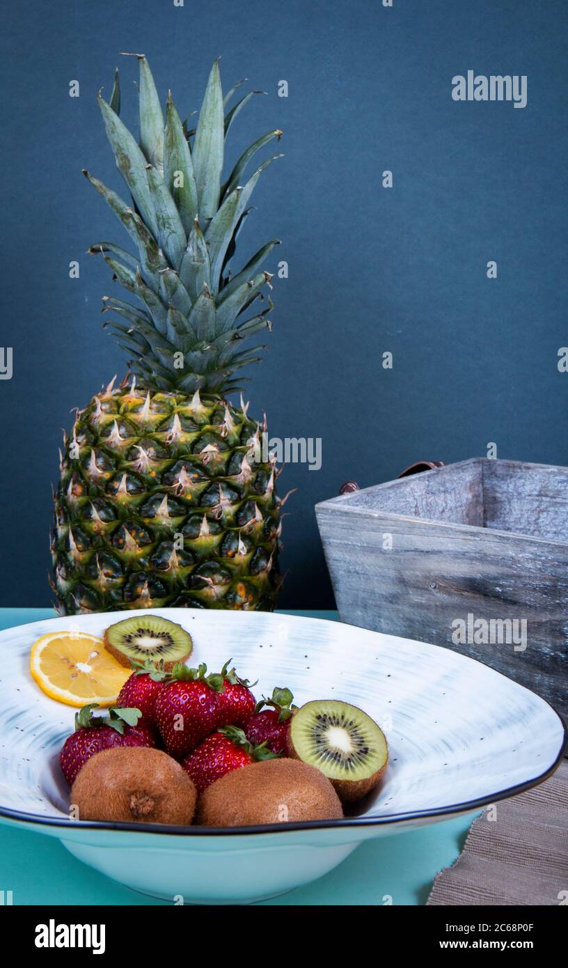 Kiwifruits, strawberries, lemon and orange section in ceramic dish and a pineapple in front of a gray wall. Stock Photo