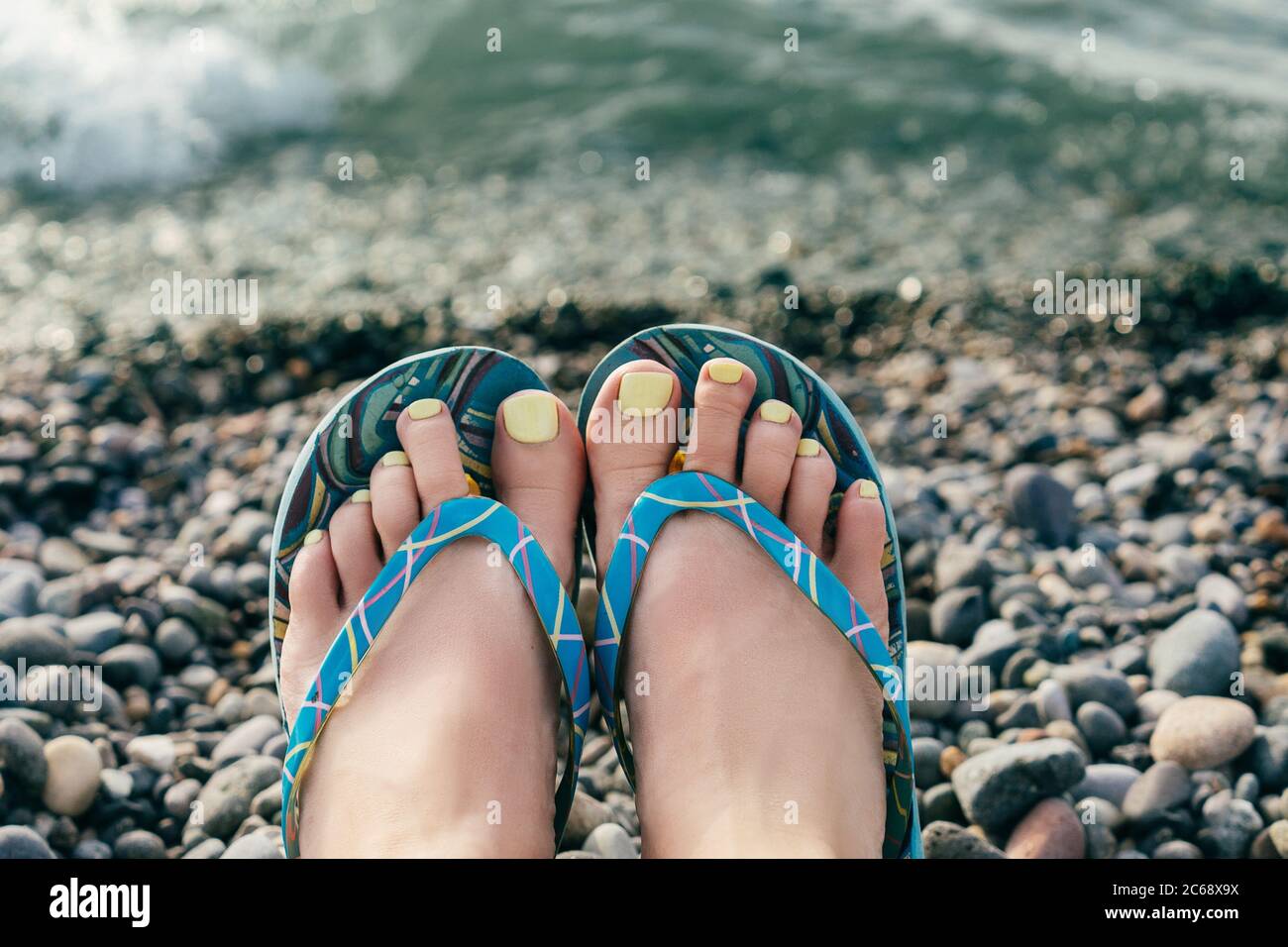 https://c8.alamy.com/comp/2C68X9X/female-legs-in-blue-flip-flops-shoes-with-perfect-yellow-pedicure-on-the-stones-beach-top-view-2C68X9X.jpg