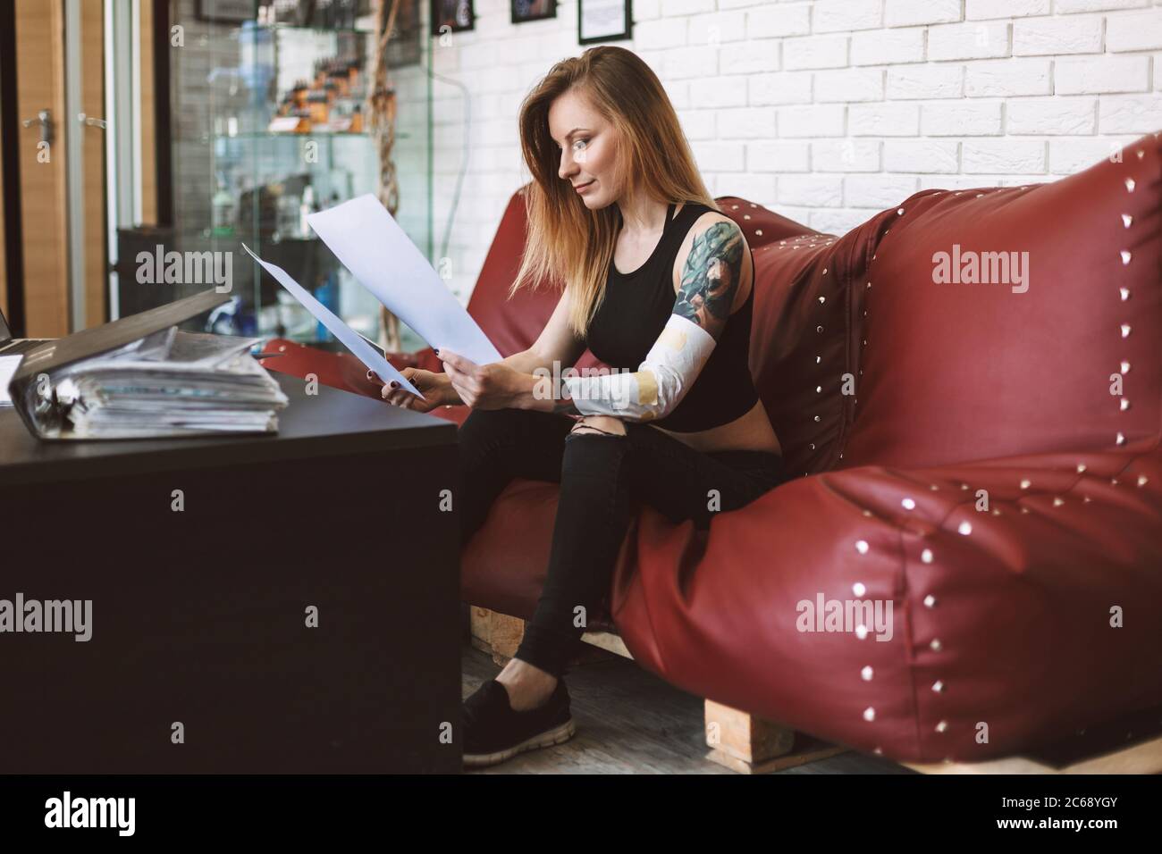Tattooed girl sitting on sofa thoughtfully watching new tattoo sketches