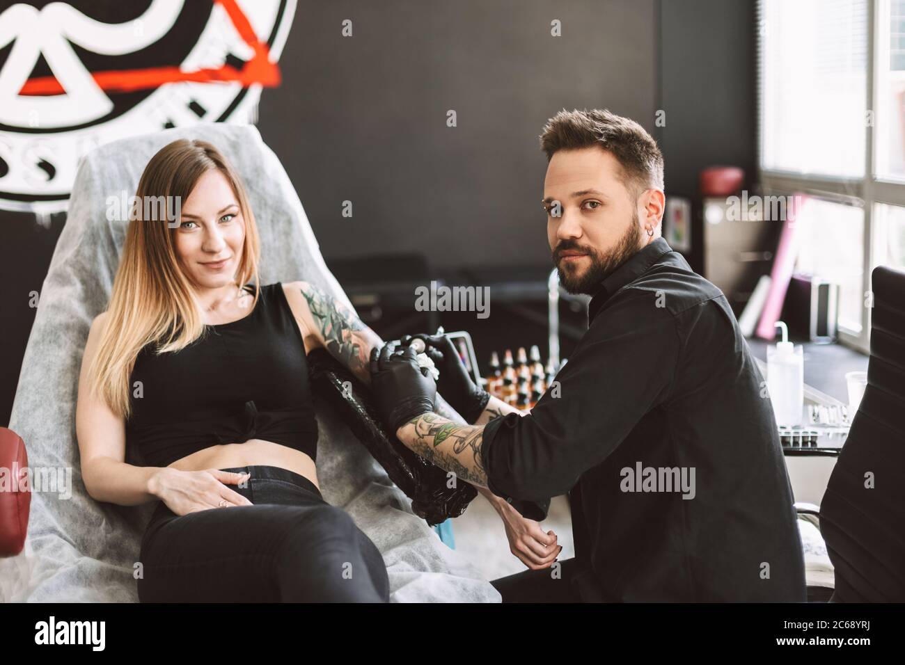 Tattooed girl and professional tattooer happily looking in camera doing tattoo on hand by tattoo machine in studio Stock Photo