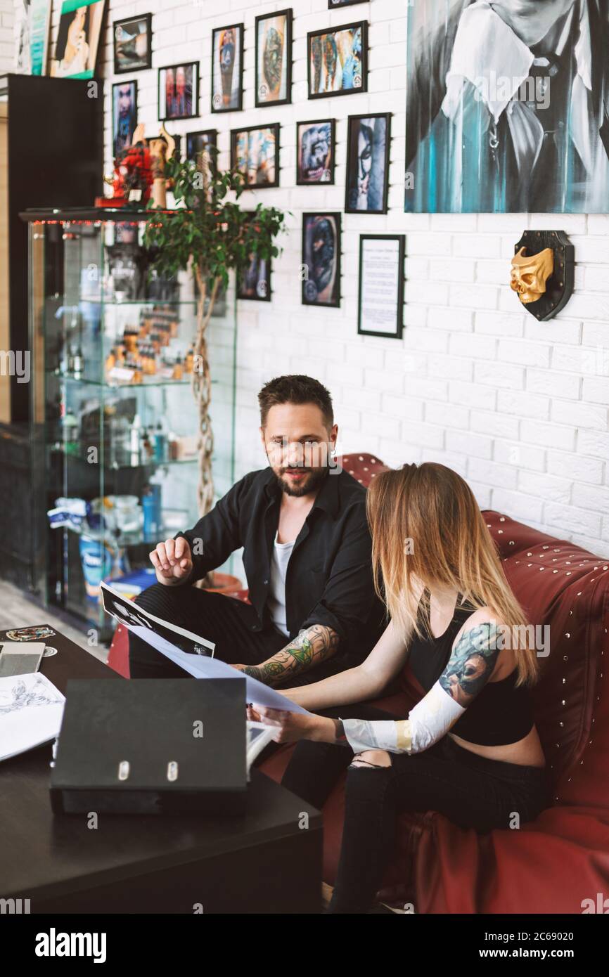 Professional tattoo master sitting on sofa with girl discussing sketch