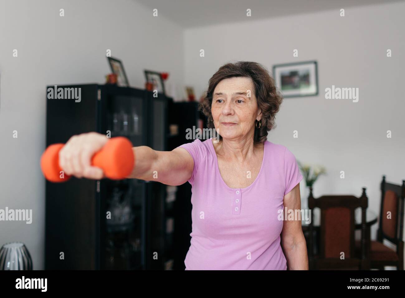 Active senior woman exercising at home. Waist up shot of 70 years old focused retired woman lifting dumbbell with her right arm in living room. Stock Photo
