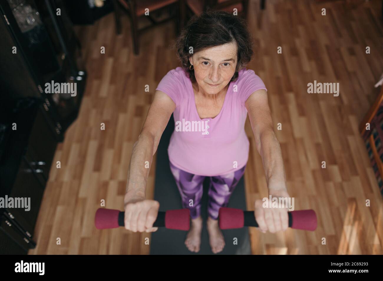 Retired woman exercising with dumbbells at home. High angle shot of 70 years old woman in purple sportswear exercising shoulders holding dumbbells bot Stock Photo